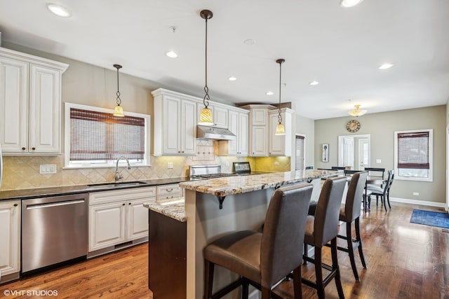 kitchen featuring a breakfast bar area, hanging light fixtures, dark stone counters, dishwasher, and under cabinet range hood