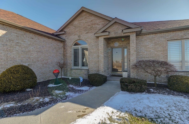 snow covered property entrance featuring roof with shingles and brick siding