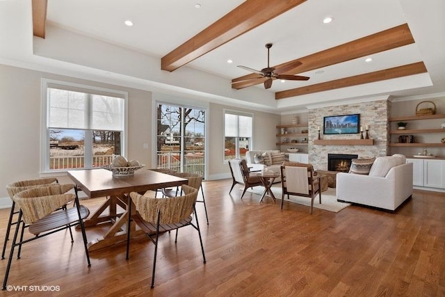 dining room with a tray ceiling, ceiling fan, dark wood-type flooring, a fireplace, and beam ceiling