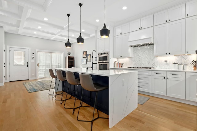 kitchen featuring light wood finished floors, white cabinets, hanging light fixtures, a kitchen island with sink, and premium range hood