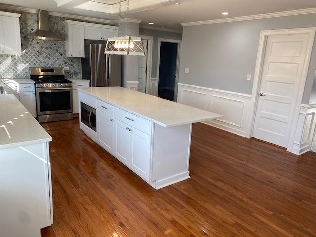 kitchen featuring appliances with stainless steel finishes, wall chimney exhaust hood, white cabinets, decorative light fixtures, and a center island