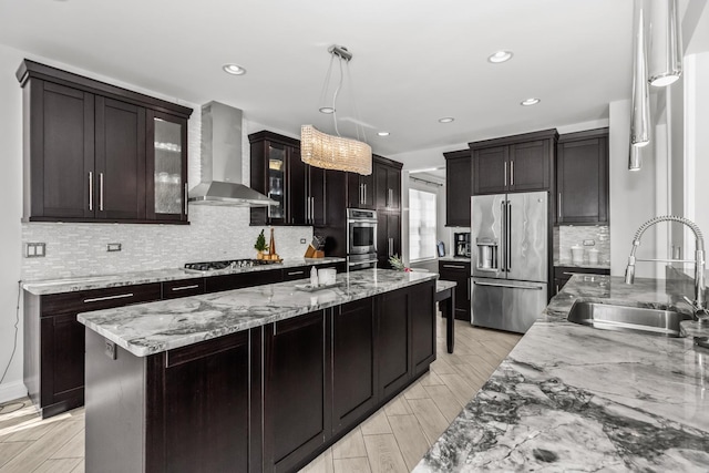 kitchen featuring a kitchen island, a sink, appliances with stainless steel finishes, wall chimney exhaust hood, and glass insert cabinets