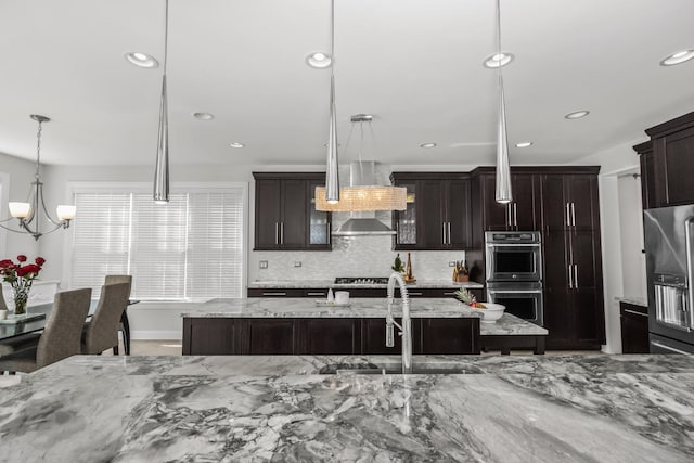 kitchen with a kitchen island with sink, a sink, hanging light fixtures, appliances with stainless steel finishes, and light stone countertops