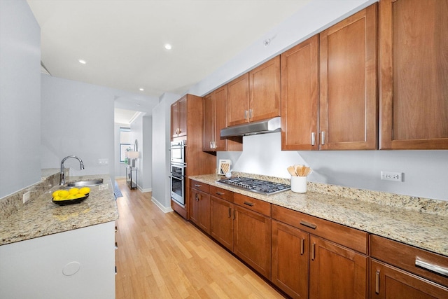 kitchen with under cabinet range hood, a sink, brown cabinets, light wood finished floors, and stainless steel gas stovetop