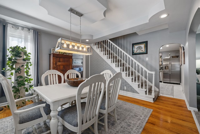 dining room featuring arched walkways, a raised ceiling, wood finished floors, stairs, and recessed lighting