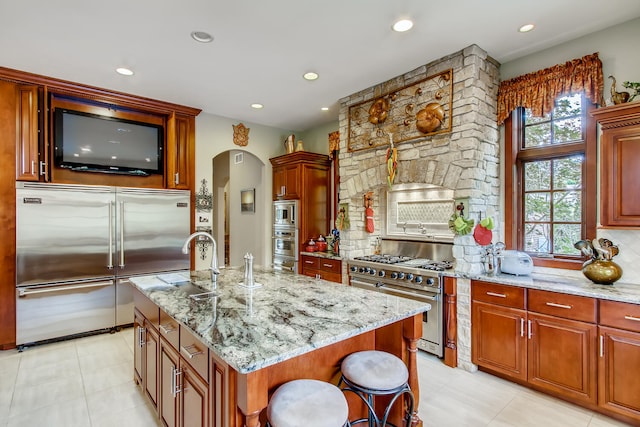 kitchen with a center island with sink, built in appliances, a breakfast bar area, light stone counters, and light tile patterned flooring