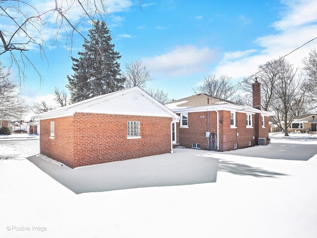 view of snowy exterior with brick siding, a chimney, and central air condition unit