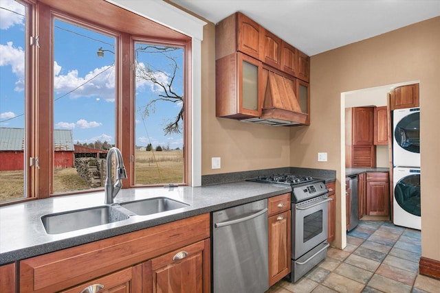 kitchen with stacked washer and dryer, a sink, dark countertops, appliances with stainless steel finishes, and brown cabinetry