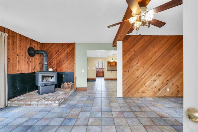 unfurnished living room featuring beamed ceiling, wooden walls, baseboards, ceiling fan, and a wood stove