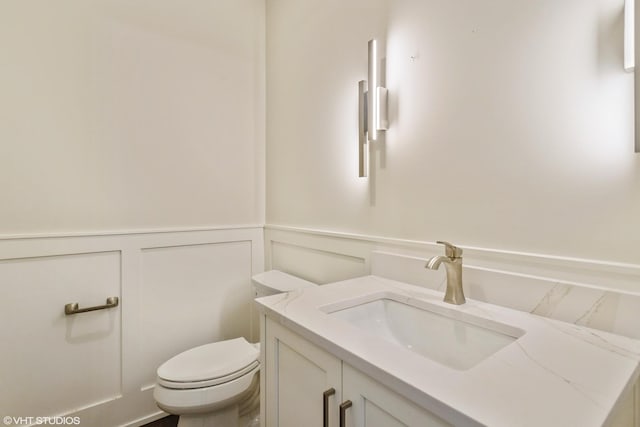 half bath with a wainscoted wall, a decorative wall, vanity, and toilet