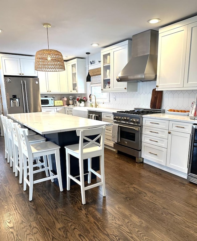 kitchen with a center island, hanging light fixtures, appliances with stainless steel finishes, glass insert cabinets, and wall chimney exhaust hood