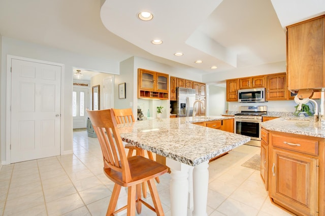 kitchen featuring light stone counters, stainless steel appliances, glass insert cabinets, a sink, and a kitchen bar