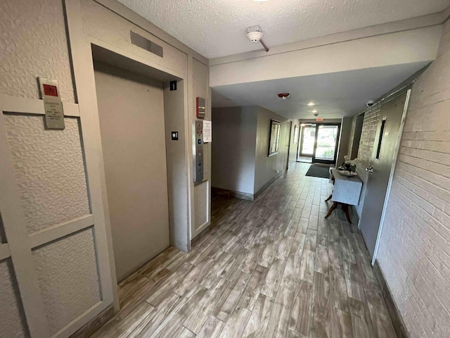 corridor featuring a textured wall, elevator, brick wall, wood finished floors, and a textured ceiling