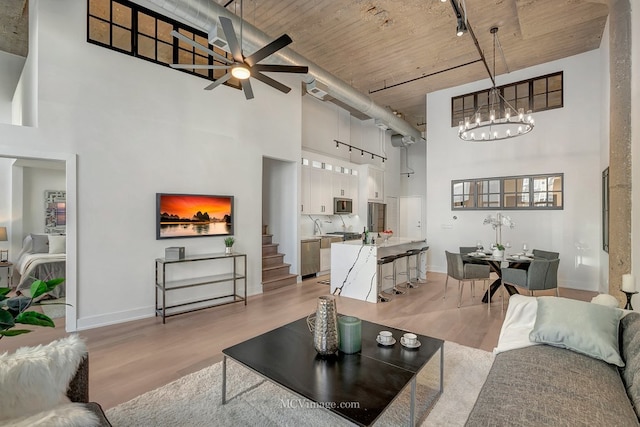 living room featuring rail lighting, light wood-style flooring, a towering ceiling, baseboards, and ceiling fan with notable chandelier