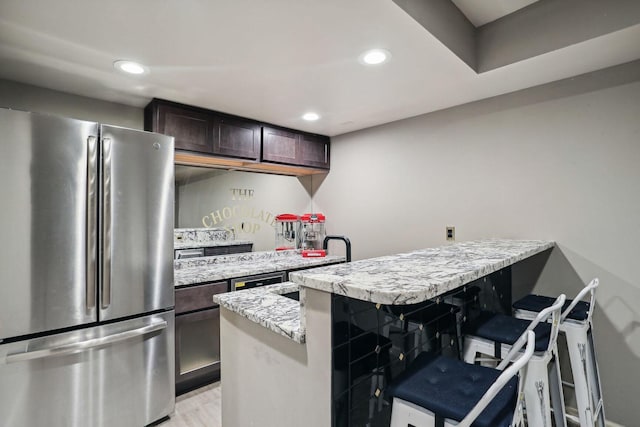 kitchen featuring light stone counters, dark brown cabinetry, recessed lighting, freestanding refrigerator, and a kitchen bar