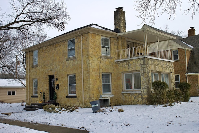 snow covered house featuring a balcony, stone siding, and a chimney