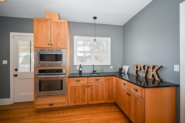 kitchen featuring light wood finished floors, dark stone counters, appliances with stainless steel finishes, decorative light fixtures, and a sink