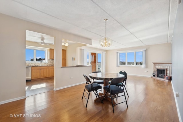 dining space with baseboards, light wood-style flooring, a textured ceiling, a brick fireplace, and ceiling fan with notable chandelier