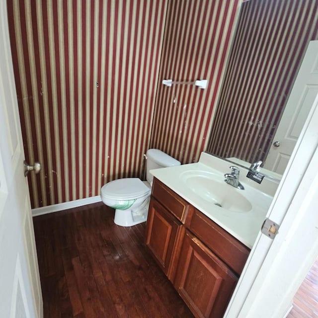 half bathroom featuring toilet, vanity, wood finished floors, baseboards, and wallpapered walls