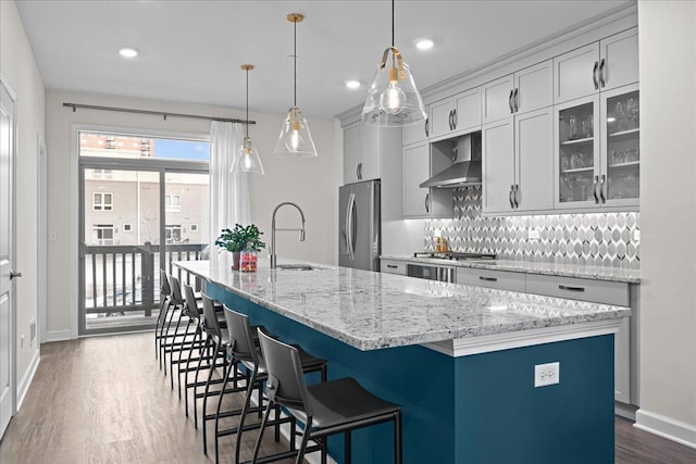 kitchen with dark wood-style floors, stainless steel appliances, tasteful backsplash, wall chimney range hood, and an island with sink