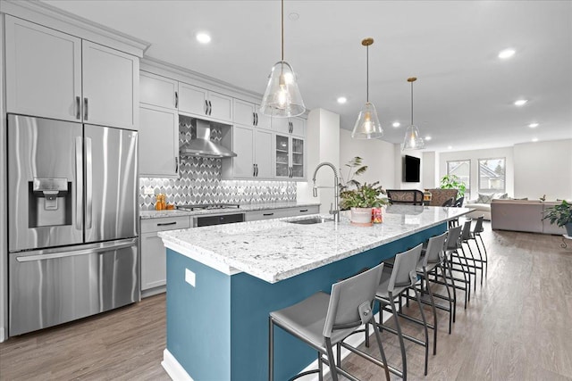 kitchen with stainless steel appliances, tasteful backsplash, a sink, wood finished floors, and wall chimney exhaust hood