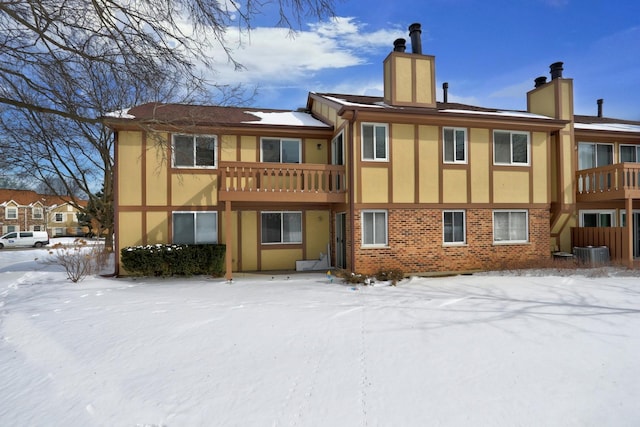 snow covered property with cooling unit, a balcony, brick siding, stucco siding, and a chimney