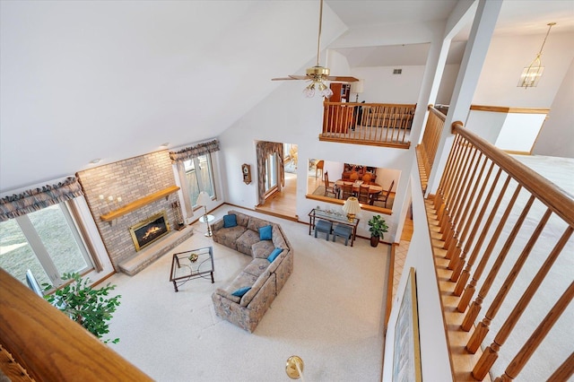 carpeted living room featuring stairs, high vaulted ceiling, ceiling fan with notable chandelier, and a brick fireplace