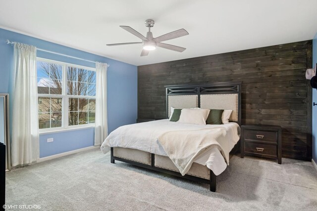 bedroom with wooden walls, light colored carpet, an accent wall, a ceiling fan, and baseboards