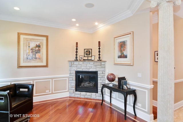 living room with a stone fireplace, a decorative wall, wood finished floors, ornamental molding, and ornate columns