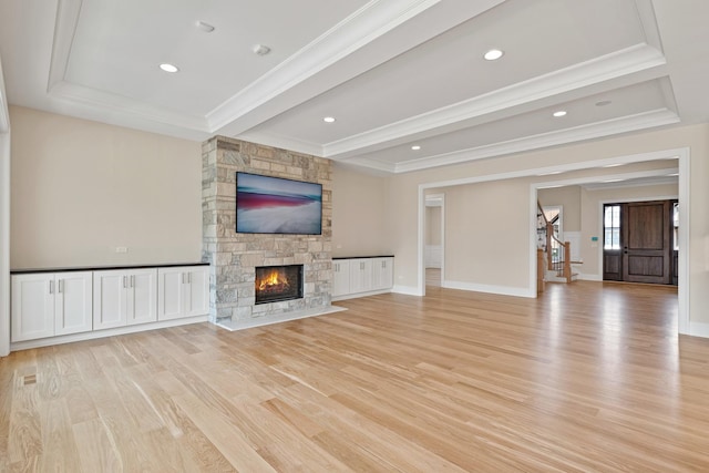 unfurnished living room featuring a tray ceiling, crown molding, light wood-style floors, a stone fireplace, and stairs