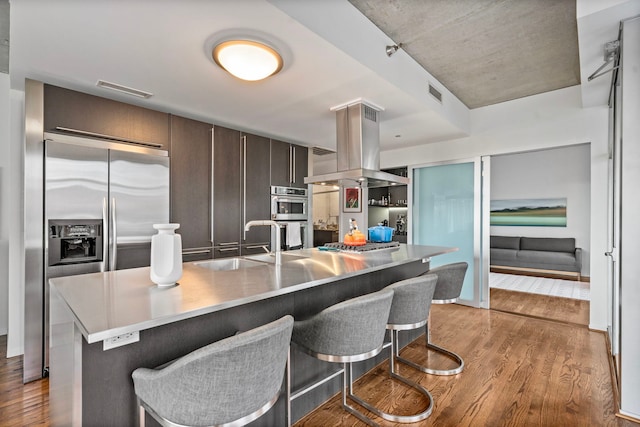 kitchen featuring dark brown cabinetry, light wood-style flooring, island range hood, a sink, and appliances with stainless steel finishes