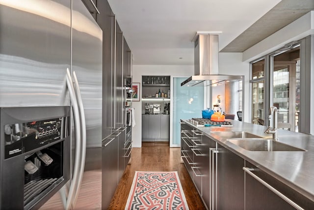 kitchen with island range hood, dark wood finished floors, stainless steel appliances, stainless steel counters, and a sink