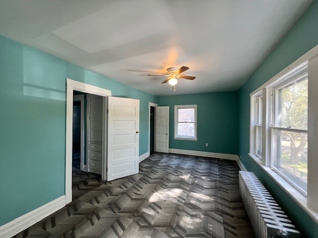 unfurnished bedroom with radiator heating unit, dark carpet, baseboards, and ceiling fan