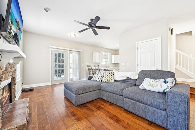 living room featuring dark wood-style floors, ceiling fan, a stone fireplace, and baseboards