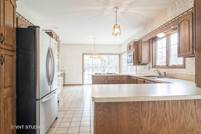 kitchen with brown cabinetry, freestanding refrigerator, a sink, a peninsula, and wallpapered walls