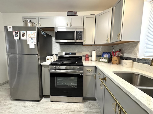 kitchen with light stone counters, appliances with stainless steel finishes, a sink, gray cabinetry, and backsplash