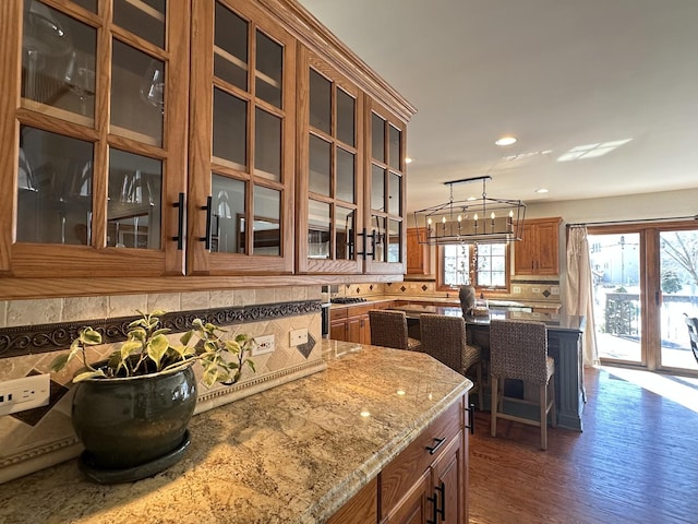 kitchen with tasteful backsplash, brown cabinetry, glass insert cabinets, dark wood-style flooring, and light stone countertops