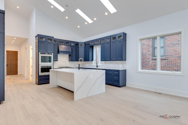 kitchen featuring an island with sink, a sink, range hood, stainless steel double oven, and a skylight