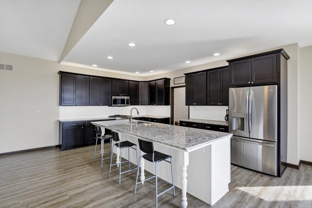 kitchen featuring stainless steel appliances, light stone counters, a center island with sink, and light wood-style floors