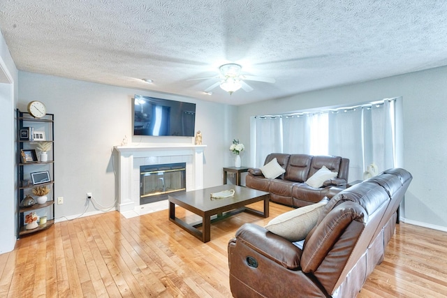 living room featuring ceiling fan, a textured ceiling, baseboards, light wood-style floors, and a tiled fireplace