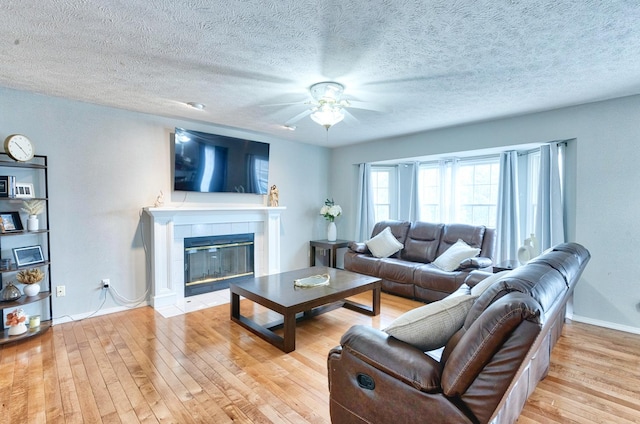 living area featuring ceiling fan, a textured ceiling, baseboards, light wood-style floors, and a tiled fireplace