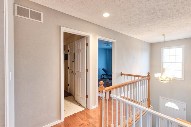 hallway with visible vents, a textured ceiling, an upstairs landing, a chandelier, and light wood-type flooring