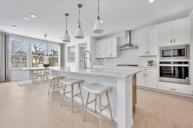 kitchen featuring wall chimney range hood, a kitchen breakfast bar, tasteful backsplash, light wood-style floors, and appliances with stainless steel finishes