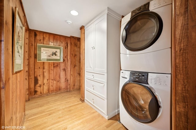 washroom featuring stacked washer and dryer, cabinet space, light wood-style flooring, and wooden walls