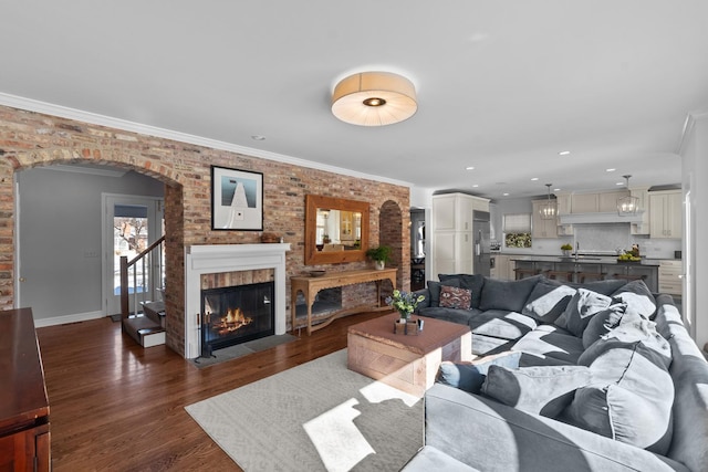 living area featuring arched walkways, brick wall, a fireplace with flush hearth, dark wood-style flooring, and crown molding