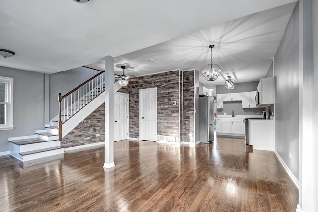 unfurnished living room with dark wood finished floors, visible vents, stairway, baseboards, and ceiling fan with notable chandelier