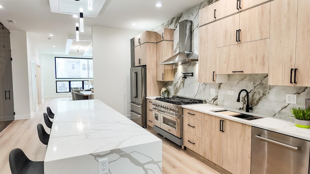 kitchen featuring premium appliances, light stone counters, light brown cabinets, a sink, and wall chimney exhaust hood