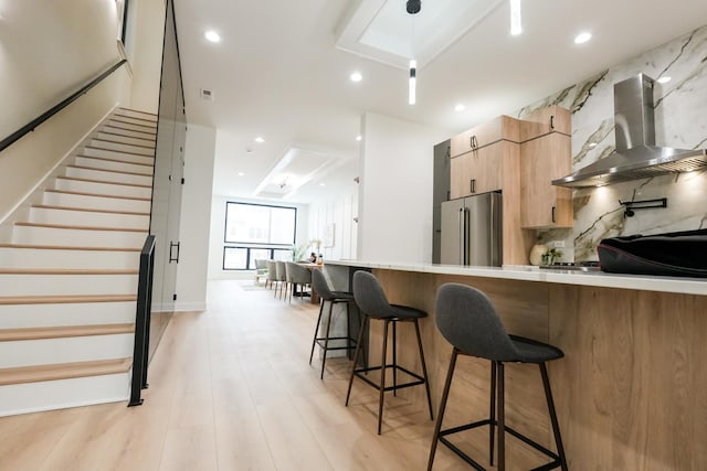 kitchen featuring high end refrigerator, wall chimney exhaust hood, modern cabinets, decorative light fixtures, and light countertops