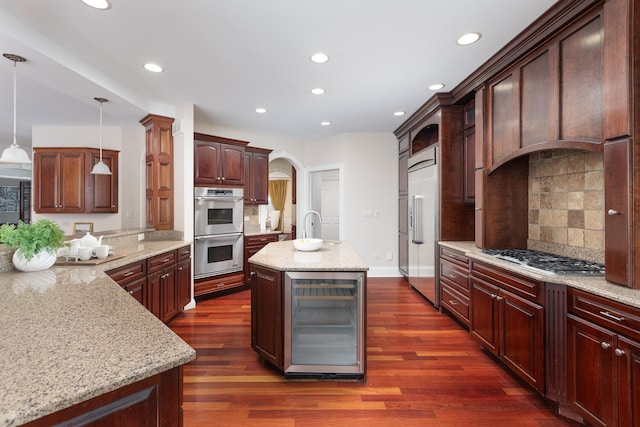 kitchen with beverage cooler, a center island with sink, arched walkways, decorative light fixtures, and stainless steel appliances