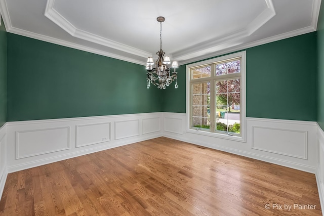 unfurnished dining area featuring a raised ceiling, wainscoting, wood finished floors, an inviting chandelier, and crown molding